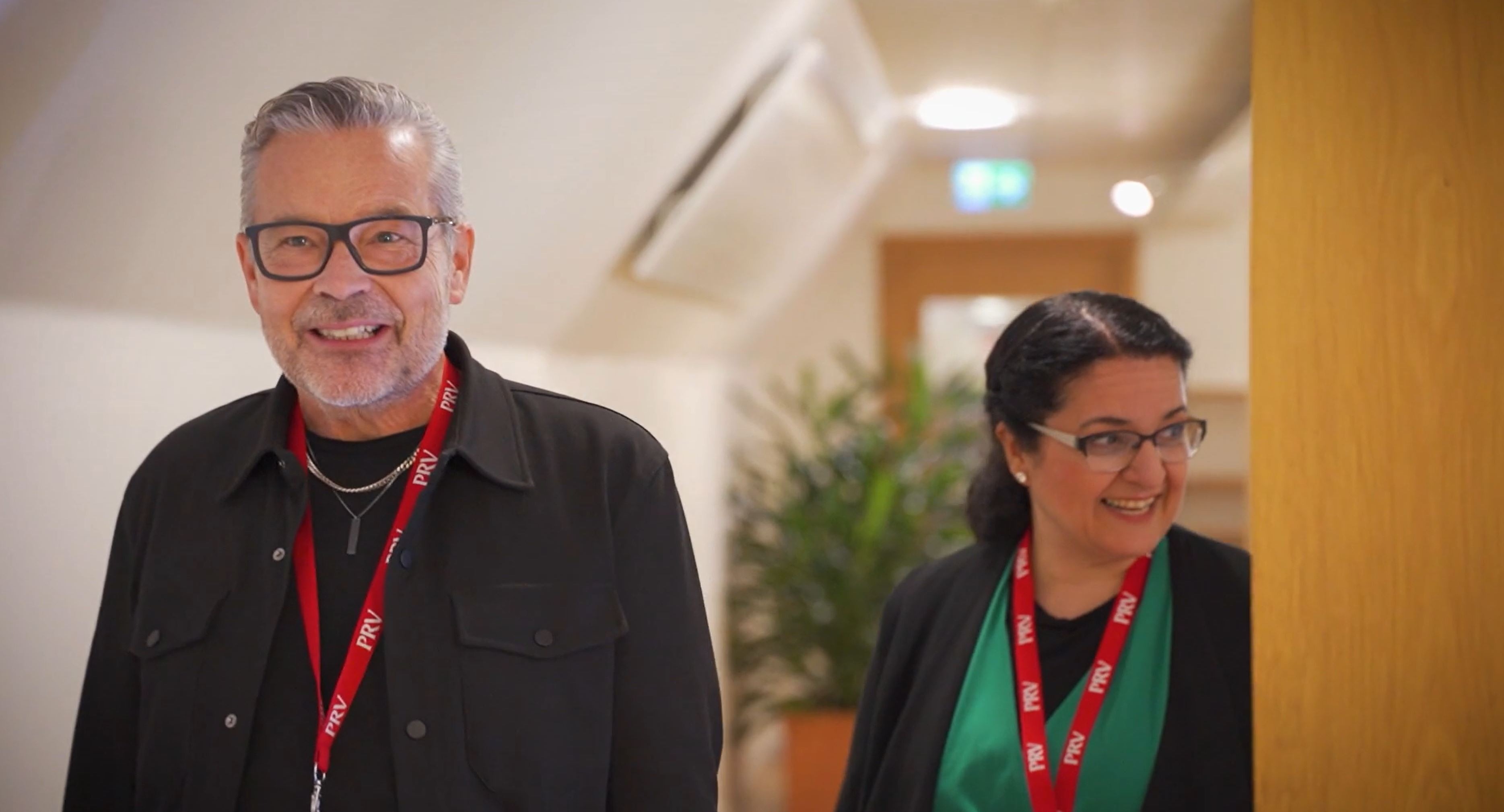 Två personer kommer gående i en korridor, de har nyckelband från PRV runt halsen. 