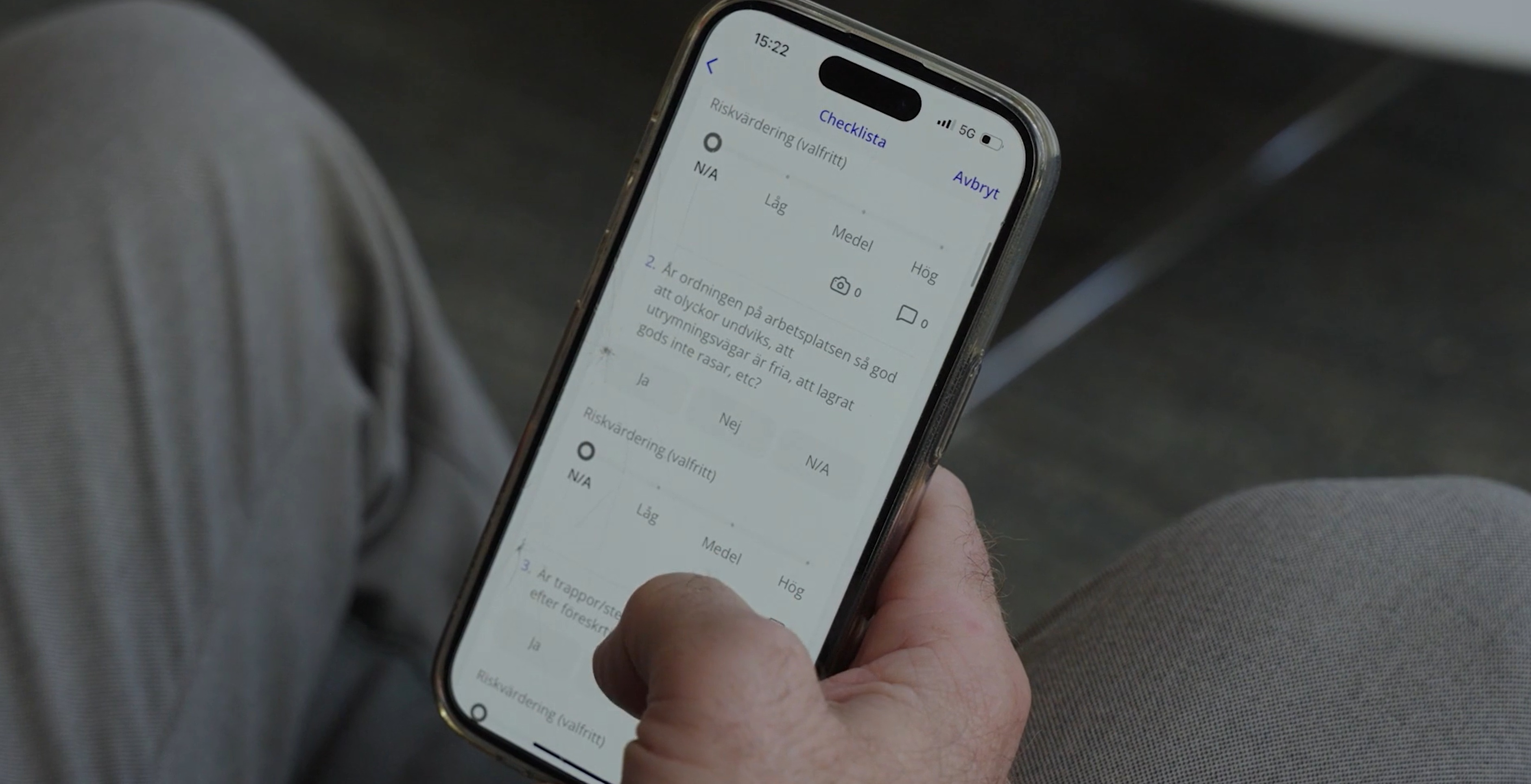 Person sitter med en mobil i handen. I mobilen syns en digital checklista från IA-systemet som kan fyllas i genom reglageknappar.